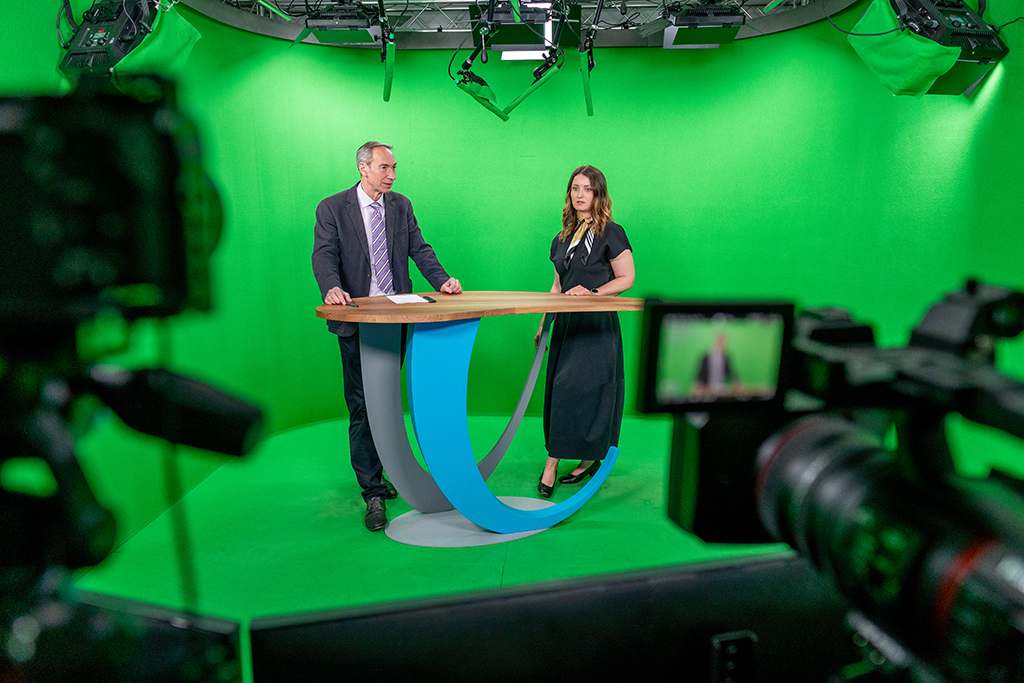 Zwei Personen stehen hinter einem modernen Studiotisch in einem komplett grünen Greenscreen-Studio. Mehrere Kameras und Studiolampen sind sichtbar. Die beiden sprechen in die Kamera, als würden sie eine Sendung oder Präsentation moderieren.