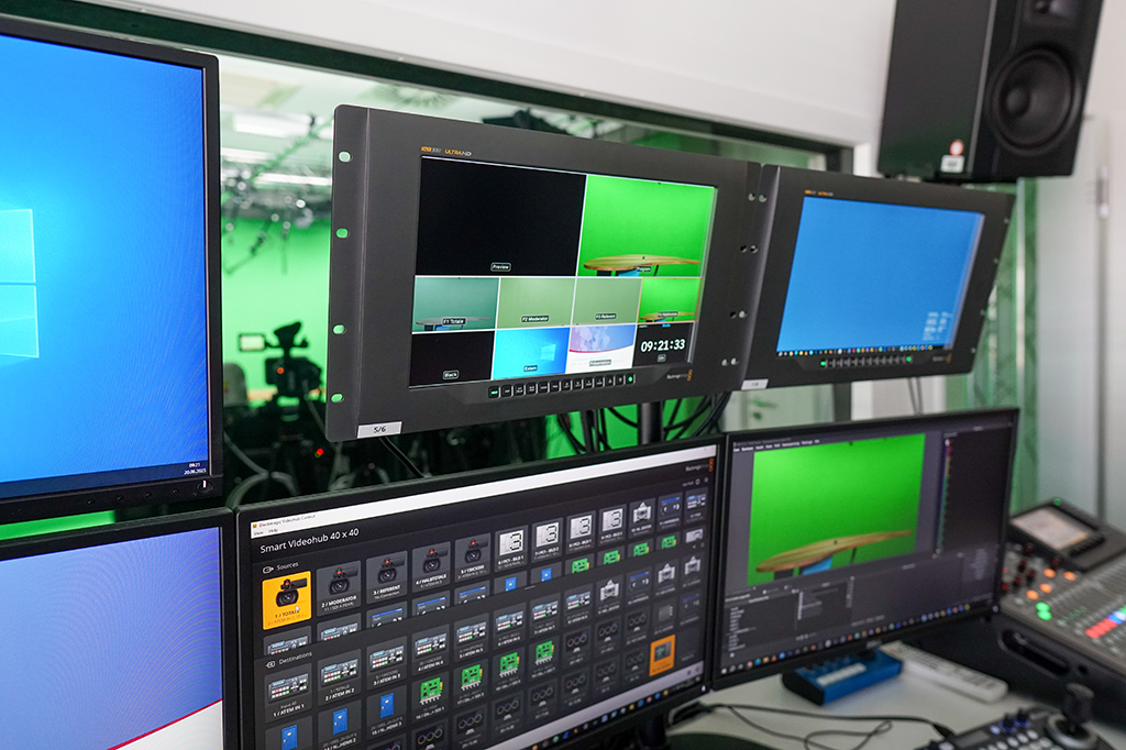Der Regieraum zeigt mehrere Monitore mit Live-Bild aus einem Greenscreen-Studio. Streaming- und Video-Software ist auf den Bildschirmen zu sehen. Audiotechnik, ein PTZ-Controller und weitere Geräte stehen auf dem Tisch.